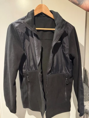 Svart fleecejacka Outdoor Grmnt - (Aldrig använd) Svart fleecejacka från Outdoor Grmnt med snygga detaljer i syntetmaterial på axlar, bröst och ärmar. Jackan har dragkedja framtill och praktiska fickor med dragkedja. Perfekt för dig som gillar outdoor och vill ha en stilren look.