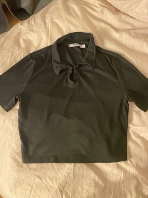 Svart croppad polotopp från NA-KD - Säljer en svart croppad polotopp från NA-KD i storlek M. Toppen har korta ärmar, krage och är tillverkad i ett ribbat material som ger en snygg struktur. Perfekt för dig som gillar stilrena och trendiga plagg.