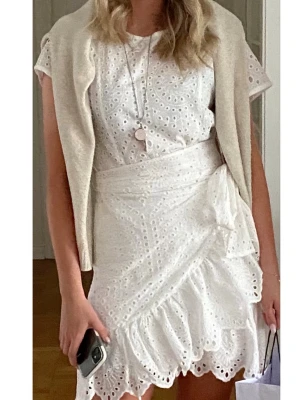 Vit omlottklänning med spetsdetaljer - Superfin vit kortklänning från By IC med broderade spetsdetaljer och omlottknytning i midjan. Klänningen har korta ärmar, rund hals och volangkant nedtill. Perfekt för sommarens alla tillfällen, riktigt söt och trendig design. Använd 1 gång.