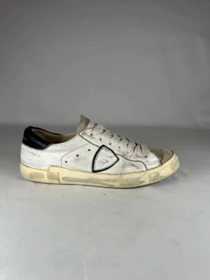 Philippe Model vita sneakers med svart häl - Snygga vita sneakers från Philippe Model med svart häl och grå mockadetaljer på tungan och tåpartiet. Skorna har snörning, rund tå och en platt sula med beige inslag. Ikonisk sköldlogga på sidan och textdetalj vid hälen. Perfekta för dig som gillar fransk streetstyle.