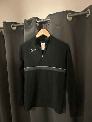 Svart Nike Dri-Fit träningströja  - Nike Dri-Fit träningströja i svart med vit logga på bröstet och en diskret grå linje över bröstet. Tröjan har half zip och är tillverkad i ett lätt syntetmaterial som andas. Perfekt för träning och sportiga aktiviteter.