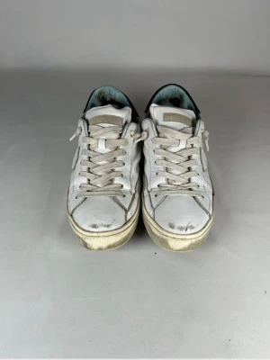 Vita sneakers från Philippe Model - Säljer ett par vita sneakers från Philippe Model med grå snörning och svart detalj på hälen. Skorna har en klassisk låg siluett, rund tå och emblem på sidan. Ovandelen är i skinn och sulan är i gummi. Perfekta för dig som gillar en clean och sportig look.