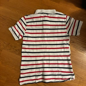 Randig vit pikétröja från Polo Ralph Lauren - Säljer en klassisk vit pikétröja från Polo Ralph Lauren med svarta och röda horisontella ränder. Tröjan har korta ärmar, krage och två knappar framtill. Den ikoniska röda broderade loggan syns på bröstet. Materialet är 100% bomull, vilket gör den mjuk och skön.