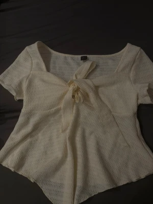 Krämvit topp med knyt från SHEIN - Säljer en krämvit topp från SHEIN i storlek M. Toppen har korta ärmar, en snygg knytning framtill och är tillverkad i ett lätt, strukturerat syntetmaterial. Den har en feminin och luftig look med rundad hals och lite loose passform.