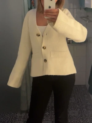 Vit kofta med knappar - Säljer en vit kofta med stora knappar och ribbade muddar. Koftan har långärmad design och två fickor framtill. Perfekt för dig som gillar en enkel och stilren look. Passar till både jeans och kjol.