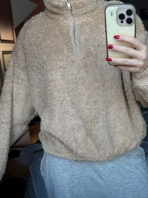 Beige teddyjacka med half zip - Supermysig teddyjacka i beige med hög krage och half zip framtill. Jackan har en loose passform och är tillverkad i fluffigt fleece-material som är perfekt när du vill vara extra cozy. Långärmad och enkel att dra på över en hoodie eller t-shirt.