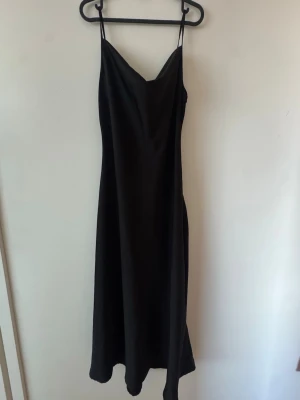 Svart lång klänning med tunna band - Säljer en svart långklänning i slip-modell med tunna axelband och mjukt, glansigt tyg. Finns mindre dragmärkeb i tyget på baksidan. Se bild
