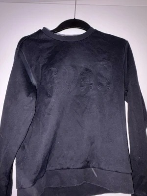 Svart Hugo Boss sweatshirt slim fit - Svart sweatshirt från Hugo Boss med stort, ton-i-ton broderat BOSS-logo på bröstet. Slim fit-modell med rund hals och ribbade muddar vid ärmslut och nederkant. Materialet är mjuk bomull som känns skön mot huden. Perfekt för dig som gillar stilrena och klassiska plagg.