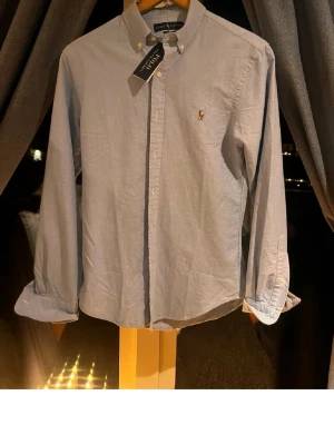 Blå skjorta från Ralph Lauren - Snygg blå skjorta från Ralph Lauren med klassisk button-down krage och broderad logga på bröstet. Skjortan har långa ärmar och knappar framtill. Perfekt för dig som gillar stilrena och tidlösa plagg.