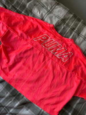 Rosa croppad t-shirt från Puma - Säljer en snygg croppad t-shirt från Puma i en stark rosa färg med diskreta ränder. T-shirten har korta ärmar och stort Puma-tryck på ryggen samt mindre logga framtill. Perfekt för dig som gillar sportig och trendig stil.