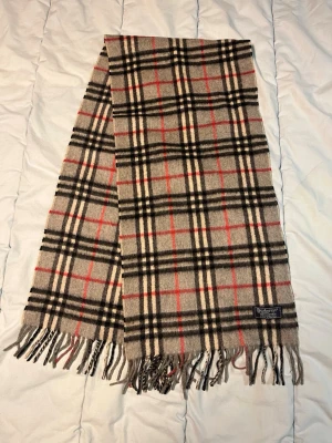 Rutig grå cashmere halsduk Burberrys - Säljer en klassisk halsduk från Burberrys i 100% cashmere. Beige botten med rutigt mönster i svart, vitt och rött. Mjukt material och snygga fransar i ändarna. Perfekt för att ge din outfit en lyxig touch. N12