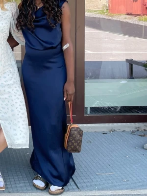 Marinblå långklänning från VILA - Säljer en elegant marinblå långklänning från VILA i storlek 34. Klänningen har en mjuk, glansig yta och ett snyggt draperat liv. Den är ärmlös och har en figurnära passform som ger en stilren siluett. Perfekt för dig som gillar klassisk och stilfull design.