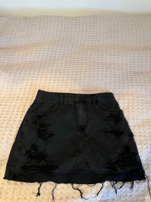 Svart jeanskjol från Hollister - Säljer en svart high-rise jeanskjol från Hollister med slitna detaljer och fransig nederkant. Kjolen har klassiska fickor fram och bak samt bälteshällor. Perfekt för dig som gillar en edgy och avslappnad stil.