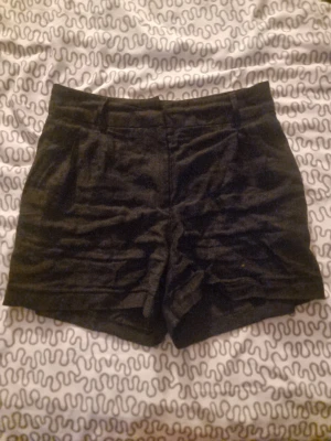 Svarta högmidjade shorts i linne - Snygga svarta shorts med hög midja och klassisk passform. Gjorda i linne för en skön och luftig känsla. Perfekta för varma dagar och har en stilren look med diskreta fickor och bälteshällor. Passar dig som gillar enkel och clean stil.