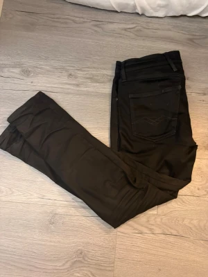 Svarta Replay Anbass jeans - Säljer ett par svarta Replay Anbass jeans med slim passform och klassisk femficksdesign. Byxorna har dragkedja och knapp framtill, samt snygga detaljer på bakfickorna. Materialet är mjukt och stretchigt för extra komfort. Perfekta för dig som gillar stilrena och moderna jeans.