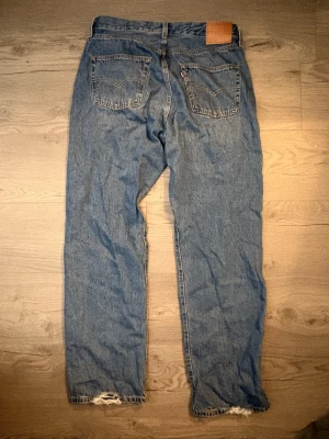 Levi's 501 jeans från 90-talet - Säljer ett par klassiska blå Levi's 501 jeans från 90-talet. De har rak passform, fem fickor och knappgylf. Jeansen är i jeansmaterial med slitna detaljer vid bensluten för en cool vintagekänsla. Perfekt för dig som gillar retrostil och tidlös denim.