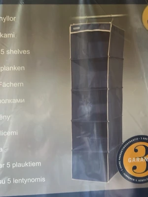 Förvaring  - Bilderna visar en förvaringshylla i tyg med fem fack, inte en väst. Produkten är mörkblå med ljusa kanter och mäter 34x34x123 cm. Ingen väst finns att beskriva.