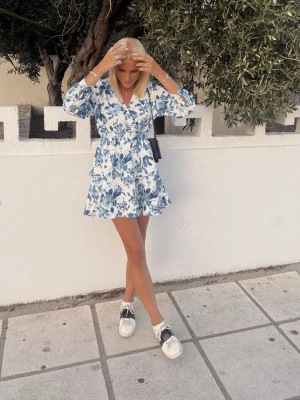 Blommig omlottklänning i blått och vitt - Superfin kort omlottklänning med blått blommönster på vit botten. Klänningen har långa ärmar, markerad midja och volangdetaljer i kjolen. Perfekt för sommarens alla tillfällen och ger en fräsch och trendig look.
