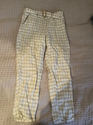 Beige-rutiga linnebyxor från Mango - Säljer ett par beige och vita rutiga linnebyxor från Mango i storlek S. Byxorna har hög midja, bälte med träspänne och avslappnad passform. Perfekta för varma dagar och ger en chill vibe till din outfit.