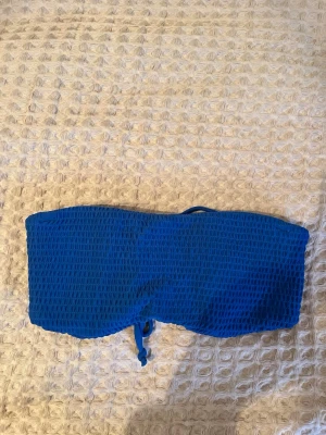 Blå bandeau bikiniöverdel Hollister - Säljer en blå bandeau bikiniöverdel från Hollister i storlek XS. Överdelen har en smockad, texturerad framsida och snörning i ryggen för justerbar passform. Perfekt för dig som gillar stilrena badkläder med coola detaljer.