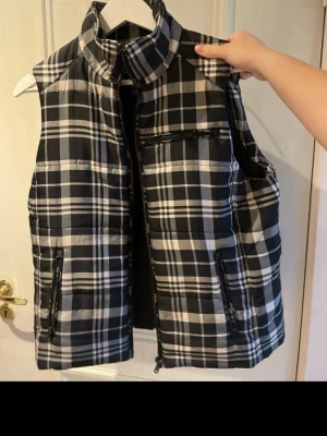 Rutig dunväst från Ralph Lauren - Säljer en snygg rutig dunväst från Lauren Ralph Lauren i storlek L. Västen har svart, vit och grå färg, hög krage och dragkedja framtill samt praktiska fickor med dragkedja. Materialet är polyester och den har en quiltad look som ger extra stilpoäng.
