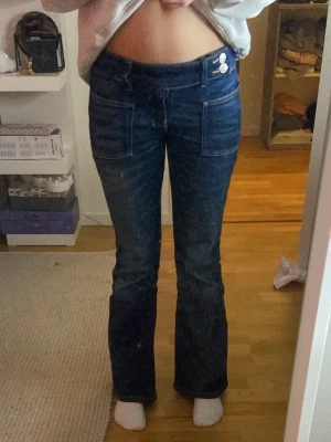 Bootcut jeans från Gina Tricot - Mörkblå bootcut jeans från Gina Tricot med två framfickor och dubbla knappar i midjan. Snygg passform med lite utsvängda ben och klassiska jeanssömmar. Perfekta för dig som gillar retrovibbar och vill ha en cool look.