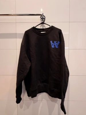 Svart sweatshirt Double A by Wood Wood - Svart sweatshirt från Double A by Wood Wood med blått tryck på bröstet och ryggen. Tröjan har rund hals, loose passform och är tillverkad i mjuk bomull. Perfekt för dig som gillar streetwear och snygga detaljer. Stora i storleken jag är 180cm och den passar. Nypris ca 1000kr, säljes för 300kr