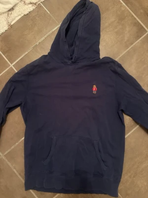 Marinblå hoodie från Polo Ralph Lauren - Snygg marinblå hoodie från Polo Ralph Lauren med klassisk huva och känguruficka. På bröstet finns den ikoniska björnen med röd tröja broderad. Tillverkad i mjuk bomull med strukturerat innerfoder i huvan. Perfekt för en avslappnad och stilren look.