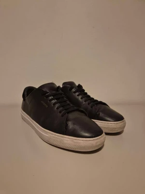 Svarta sneakers från Axel Arigato - Stilrena svarta sneakers från Axel Arigato i slätt skinn med klassisk låg profil och vit sula. Diskret logga i guld på sidan och svarta snören. Perfekta för en clean och modern look.