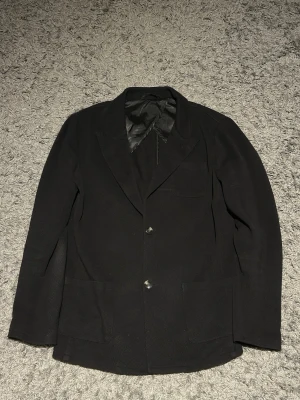 John Henric kavaj - Stilren svart kavaj med två knappar framtill, klassisk krage och tre fickor. Kavajen har en slät yta och är helfodrad för extra komfort. Perfekt för dig som vill ha en enkel men snygg look.