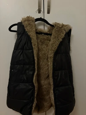 Svart dunväst med fuskpäls - Snygg svart dunväst med mjukt beige fuskpälsfoder och huva. Västen har två fickor med dragkedja och quiltad design. Perfekt för lager på lager och ger en cool look till höst- och vinteroutfits.