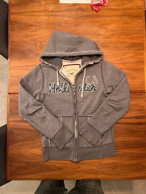 Hollister hoodie med slitningar 🥹😍 - Säljer en grå hoodie från Hollister med dragkedja och vit broderad logga på bröstet. Hoodien har vita snören i huvan, två fickor fram och ribbade muddar vid ärmar och midja. Perfekt för en avslappnad och stilren look.