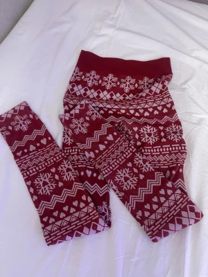 Vinröda leggings med vintermönster - Mysiga vinröda leggings med vita snöflingor, hjärtan och zigzag-mönster. Perfekta för kalla dagar och har en bred midjeresår för extra komfort. Materialet känns mjukt och stretchigt, vilket gör dem riktigt sköna att ha på sig.
