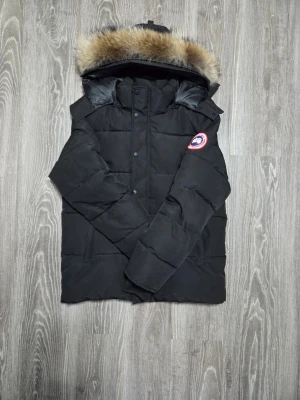 Svart dunjacka från Canada Goose - Säljer en svart dunjacka från Canada Goose med stor huva och snygg pälskant. Jackan har quiltad design, knappar och dragkedja framtill samt den klassiska loggan på ärmen. Perfekt för kalla vinterdagar och riktigt varm.