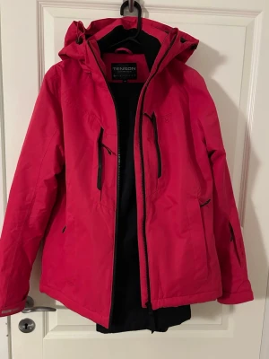 Skidjacka och byxa från Tenson - Snygg röd vindjacka (strl 40) från Tenson med justerbar huva och svarta dragkedjor. Jackan har flera praktiska fickor framtill och väldigt mjuk och skön på. Matchande skidbyxor i svart (strl 42). Mkt fint skick!