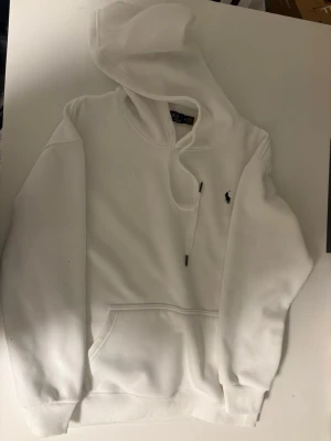 Vit hoodie från Polo Ralph Lauren - Snygg vit hoodie från Polo Ralph Lauren med klassisk broderad logga i svart på bröstet. Hoodien har huva med dragsko, magficka och ribbade muddar vid ärmar och nederkant. Tillverkad i mjukt bomullsmaterial för skön känsla. Liten i storlek så rekommenderas för Small användare. Replic hoodie