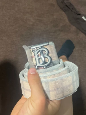 Beige rutigt bälte med metallspänne - Säljer ett beige bälte från Burberry med klassiskt rutmönster och ett stort, silverfärgat metallspänne med BB-logga. Bältet har en bred design och är tillverkat i tygmaterial med tydliga Burberry-detaljer. Perfekt för att lyfta din outfit. Helt ny 