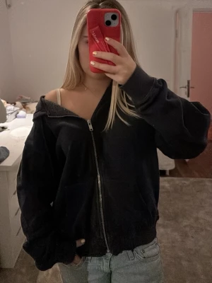 Svart zip-hoodie med huva - Säljer en svart hoodie med dragkedja och huva. Hoodien har en avslappnad passform, långa ärmar och ribbade muddar. Perfekt för en chill och casual stil. Materialet känns mjukt och bekvämt, och den har fickor framtill.