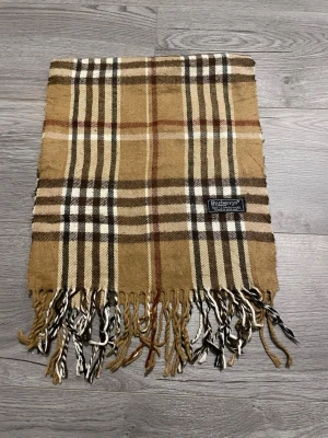  Burberrys halsduk - Säljer en ikonisk beige halsduk från Burberrys i 100% lammull. Den har det klassiska rutmönstret i beige, brunt, vitt och rött samt fransar i ändarna. Perfekt accessoar för att lyfta din outfit med en tidlös brittisk touch.