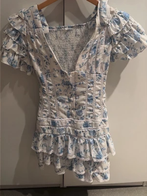 Blommig kortklänning med volanger - Supersöt kortklänning med blått blommönster på vit botten. Klänningen har volangdetaljer vid ärmar och kjol, v-ringning och smockad rygg för skön passform. Puffiga kortärmar och omlottkänsla framtill ger en romantisk vibe. Perfekt för sommarens alla tillfällen!