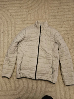 Ljusgrå dunjacka från Everest - Snygg ljusgrå dunjacka från Everest med quiltad design och svart dragkedja. Jackan har hög krage, två sidofickor med dragkedja och diskret logga på bröstet. Perfekt för kyliga dagar och enkel att matcha med olika outfits.