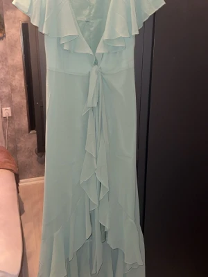 Ljusgrön långklänning med volang - Säljer en elegant ljusgrön långklänning med volangdetaljer och knyt framtill. Klänningen har v-ringning, ärmlös design med bred volang runt axlarna.