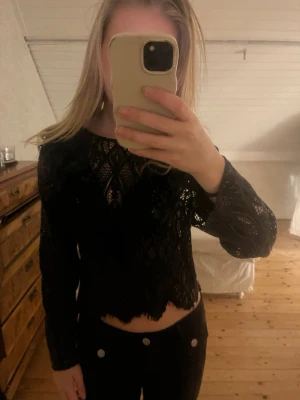 Svart spetsblus med lång ärm - Säljer en svart  blus i spets med lång ärm och rund hals. Blusen har fina spetsdetaljer och vågig kant nertill som ger en edgy look. Perfekt att styla med byxor för en trendig outfit.