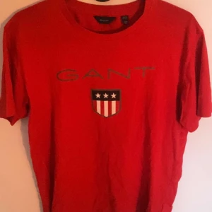 Röd GANT t-shirt  - Snygg röd t-shirt från GANT med klassiskt tryck framtill. T-shirten har korta ärmar och rund halsringning. På bröstet finns GANT-loggan och ett sköldmärke med stjärnor och ränder. Perfekt för en avslappnad och stilren look.