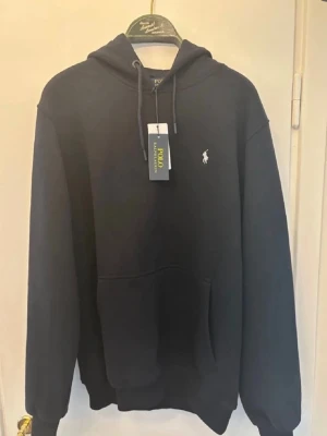 Svart hoodie från Polo Ralph Lauren - Snygg svart hoodie från Polo Ralph Lauren med klassisk huva och dragsko. Har en liten vit logga broderad på bröstet och en stor magficka framtill. Perfekt för en avslappnad och stilren look. Tillverkad i mjukt material för extra komfort.