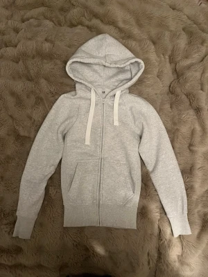 Russemerch hoodie  - Ljusgrå hoodie med dragkedja från Russell. Klassisk modell med huva och vita dragsnören, två fickor framtill och ribbade muddar vid ärmar och midja. Tillverkad i mjukt sweatshirtmaterial som är perfekt för en avslappnad stil.