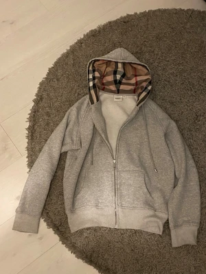 Grå zip hoodie från Burberry - Säljer en grå hoodie från Burberry med dragkedja och klassiskt rutigt foder i huvan. Hoodien har två fickor framtill, justerbar dragsko i huvan och ribbade muddar. Perfekt för dig som gillar stilrena plagg med en twist av lyx. De står storlek M men skulle defenitivt säga att den är mer S kanske till och med XS. Vid frågor eller annat hör av er