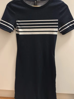 Marinblå kort klänning från H&M - Kort marinblå klänning från H&M Divided med vita horisontella ränder över bröstet och på ärmarna. Klänningen har rund halsringning och korta ärmar. Tillverkad i mjukt, stretchigt material som sitter snyggt och bekvämt.
