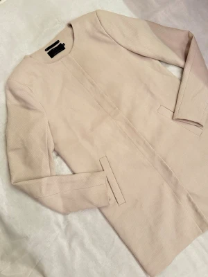 Kräm vit kappa från ONLY - Stilren lätt jacka från ONLY med rak passform och diskreta fickor framtill. Kappan har rund halsringning och långa ärmar. Materialet känns mjukt och har en lätt struktur som ger en modern look. Perfekt för dig som gillar minimalistisk stil.