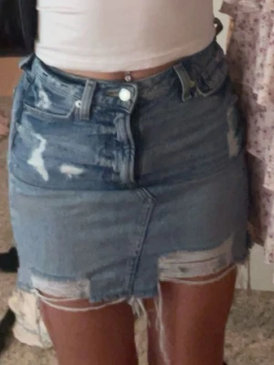 Blå jeanskjol med slitningar - Säljer en kort jeanskjol i blå denim med slitna detaljer och rå kant nertill. Kjolen har klassisk femficksdesign, hög midja och dragkedja med knapp framtill. Perfekt för dig som gillar en avslappnad och trendig stil.